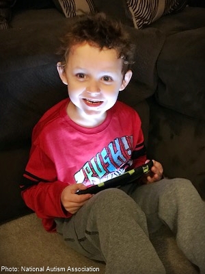 a young boy with curly hair and a red sweatshirt sitting on the floor with his tablet